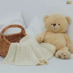 A plush teddy bear sits beside a wicker basket with a cream knitted blanket, against white pillows.