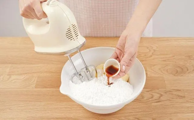 A person is using an electric mixer to prepare food on a table covered with kitchen utensils and tableware.