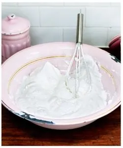 A whisk is vigorously mixing a thick paste of cream and a thickening agent in a mixing bowl on a kitchen table surrounded by dairy, kitchenware, dishware, tableware, and serveware.