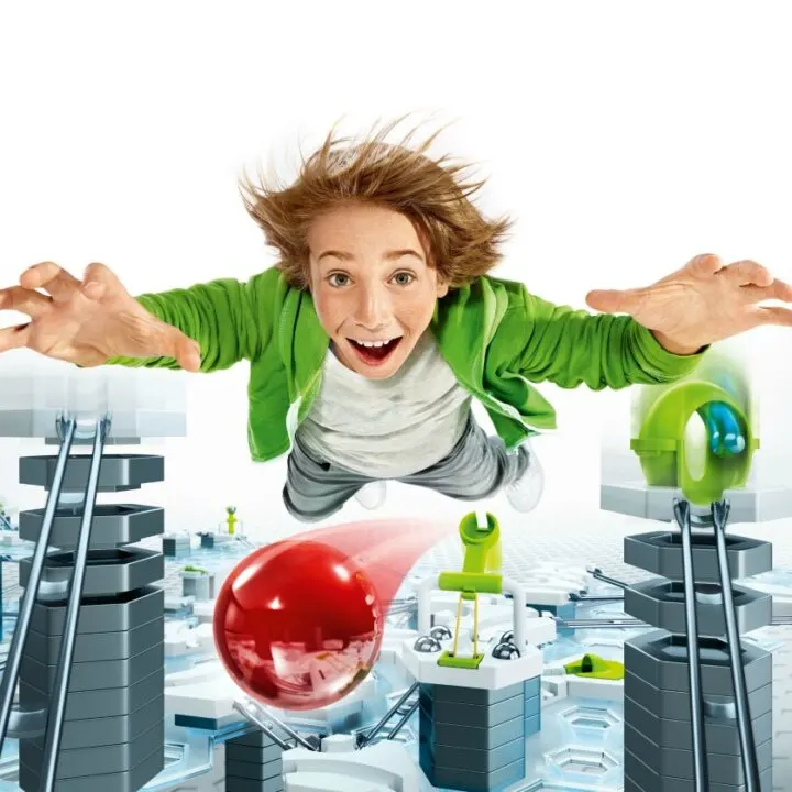 A cartoon child with a smiling face holds a balloon in one hand while playing with a GRAVITRAX set in the other.