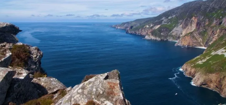 A panoramic view of a coastal landscape featuring a mountain, raised beach, bay, cliff, escarpment, cove, island, peninsula, inlet, and other oceanic landforms in the sky above a body of water.