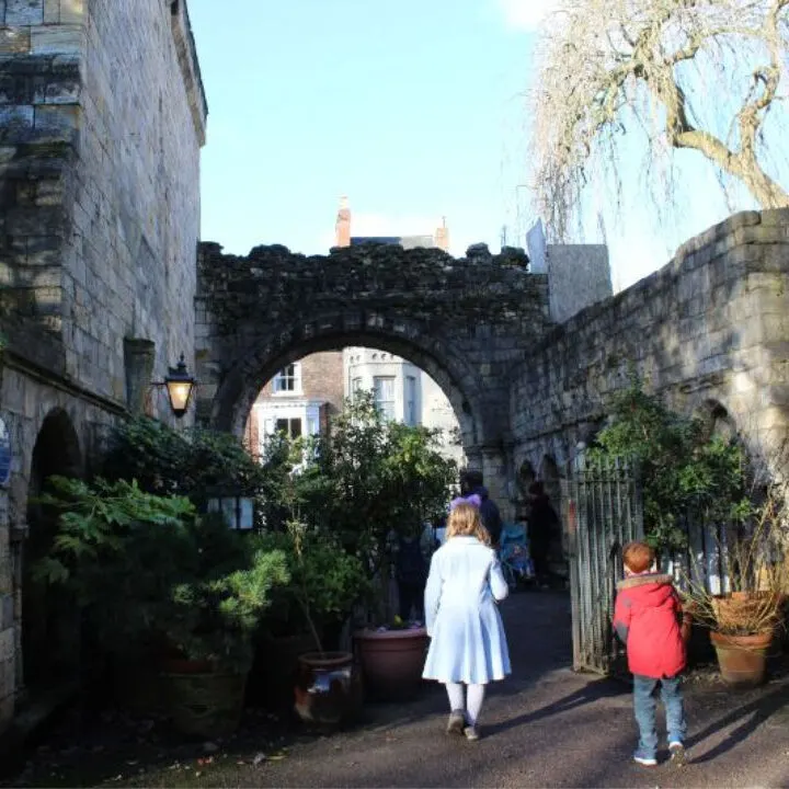 People of all ages are standing in awe of the majestic castle with its stone arch and lush plants, surrounded by a bustling city street and a clear blue sky.