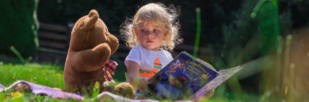 child reading a book next to her teddy bear