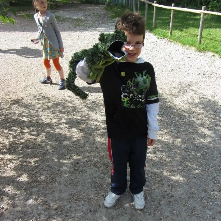 A young person stands in an outdoor playground surrounded by a tree, plant, and toddler.