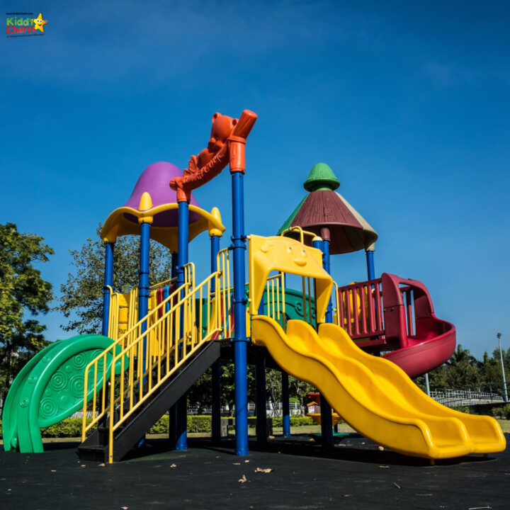 Children are playing on a yellow playground slide surrounded by outdoor play equipment, trees, and a clear blue sky in a public park.