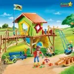 A bright blue sky dotted with fluffy white clouds provides a picturesque backdrop to a lush green tree, outdoor play equipment, and a grassy playground.