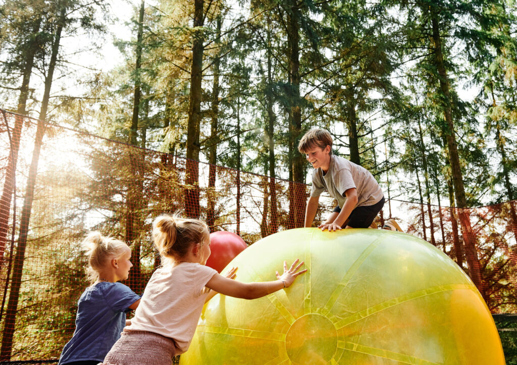 A group of people playing on a large yellow and green ball in a forest.