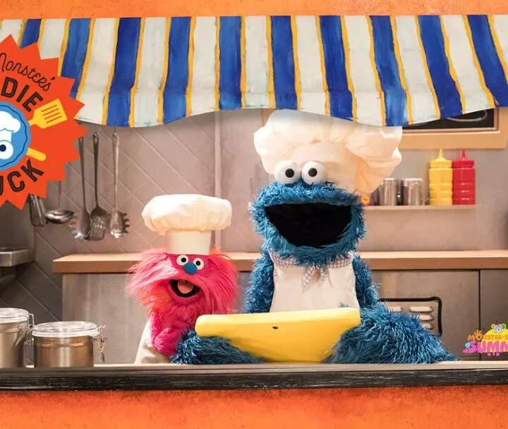 A cartoon of Cookie Monster driving a toy food truck inside a kitchen fills the frame.