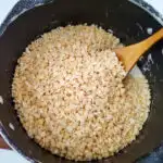 A cook is stirring a mixture of food grains in a wok, while a plate of cooked rice sits on a bowl and pan of cookware and bakeware in an indoor kitchen.