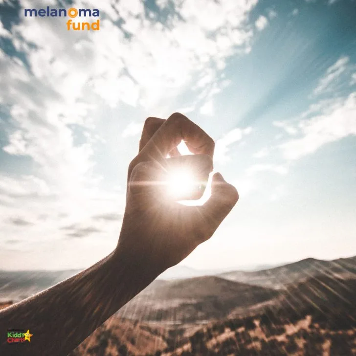A person stands in a desert landscape with a mountain in the background, looking up at a sky filled with clouds and a sun, while holding a screenshot of the Kiddy & Charts Melanoma Fund.