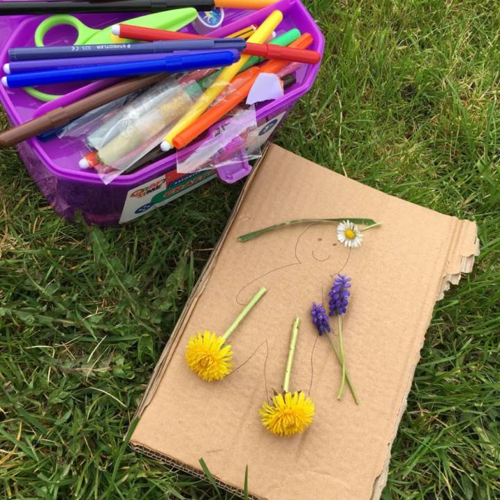 A child is creating art outdoors with a STAEDTLER 325 CE pen, scissors, and other office supplies amidst a lush grassy landscape.