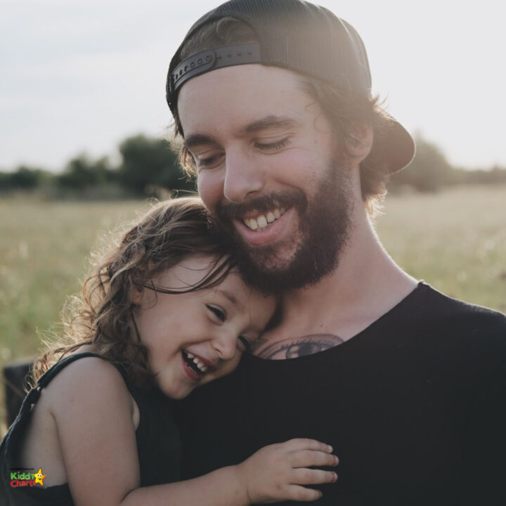 A smiling person with a beard embraces a person lovingly in a grassy outdoor setting beneath a bright blue sky.