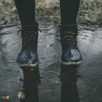 A person stands in a puddle of water, ankle-deep, wearing a pair of wet boots and shoes.