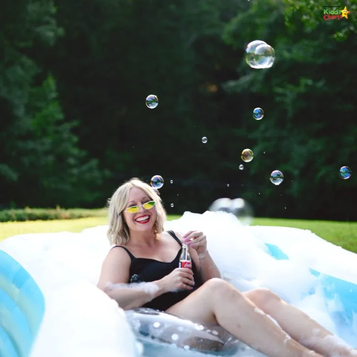 A person and a person are enjoying a summer day outdoors, with the person lying in the grass and the person standing in a pool of water beneath a tree with a human face in its trunk, both of them looking up at a bubble in the sky.