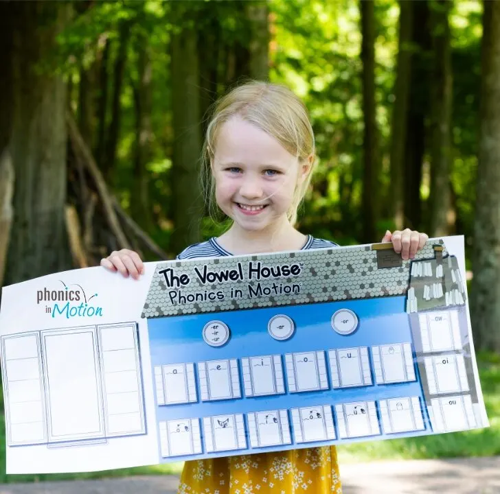 A young person smiles while standing in a park, holding a sign with text about the Vowel House in Motion Phonics in Motion Charts.