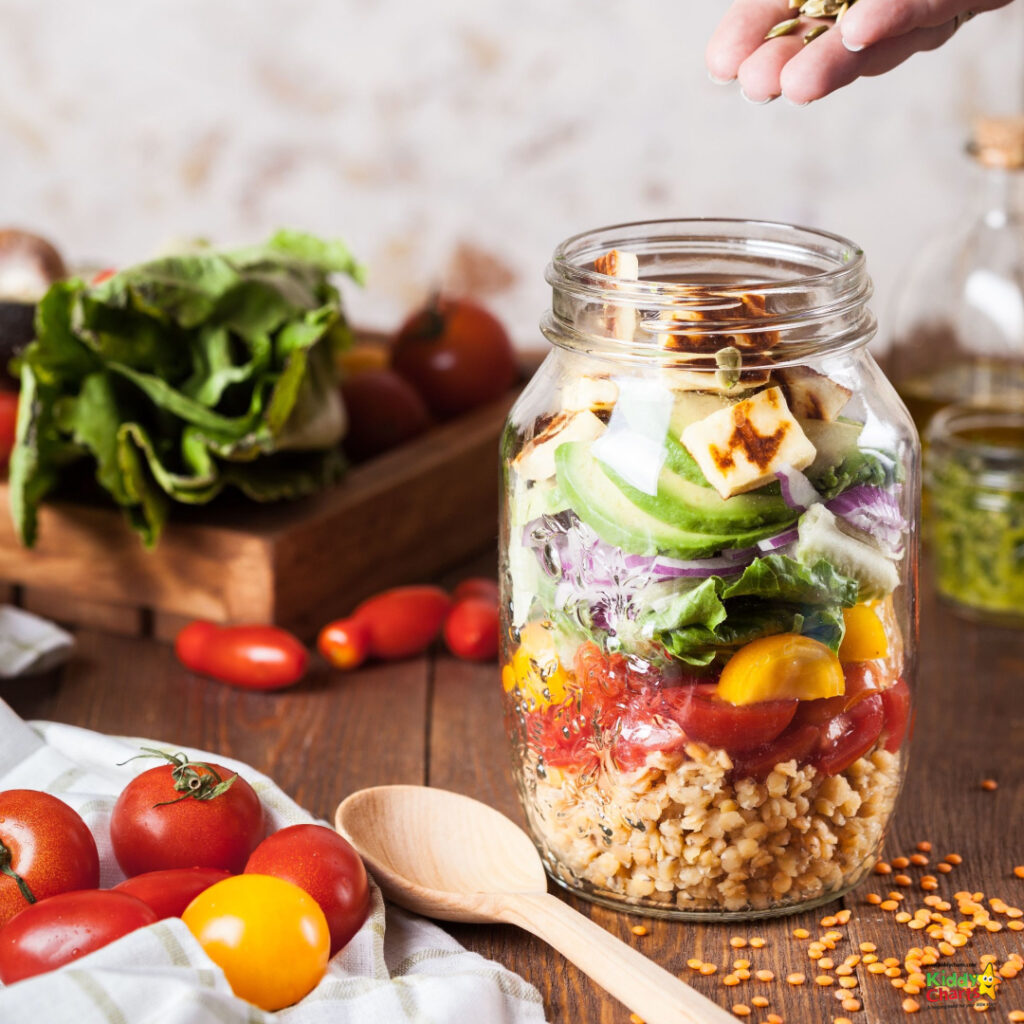 A person is cutting fresh cherry tomatoes on a table surrounded by a variety of natural foods, vegan nutrition, vegetarian food, superfoods, and staple foods, all stored in mason jars and food storage containers, to create a healthy and balanced diet.