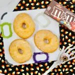 Three glazed donuts on a candy corn cloth, with a skeleton hand holding a toy eye, M&M's box, and ghost-shaped cookie cutters.