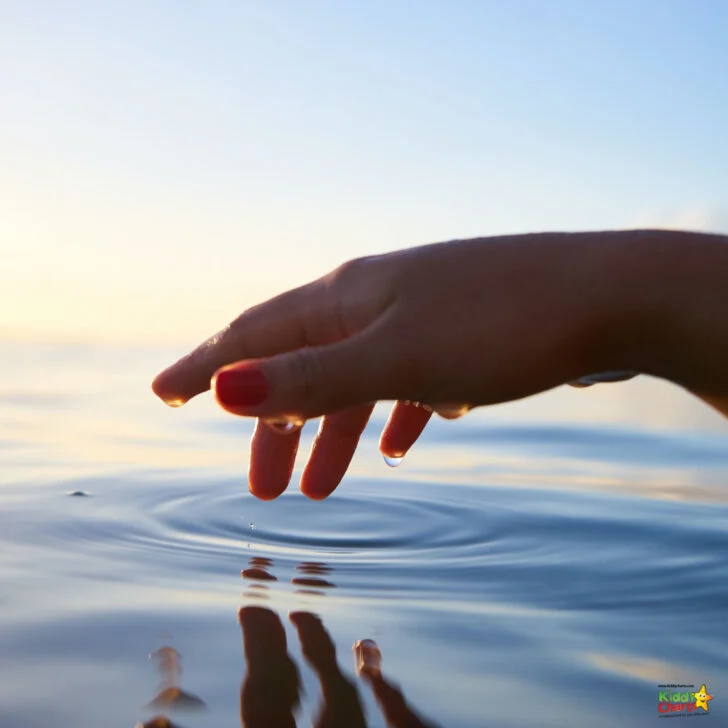 A person stands on a beach, their hand outstretched and fingers spread wide, creating a wave in the lake's fluid surface beneath a bright sky.