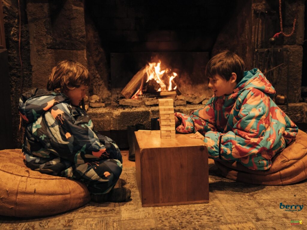 Two children in colorful clothing sit on bean bags playing Jenga near a fireplace, with a cozy and warm atmosphere.