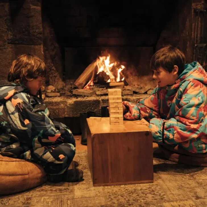 Two children in colorful clothing sit on bean bags playing Jenga near a fireplace, with a cozy and warm atmosphere.