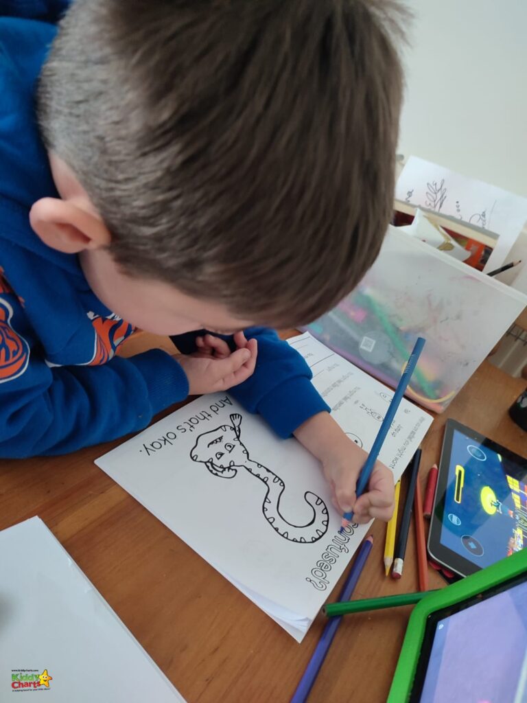 A child is intently coloring a printed outline of a cartoon snake, surrounded by colored pencils. There is a smartphone and tablet nearby.