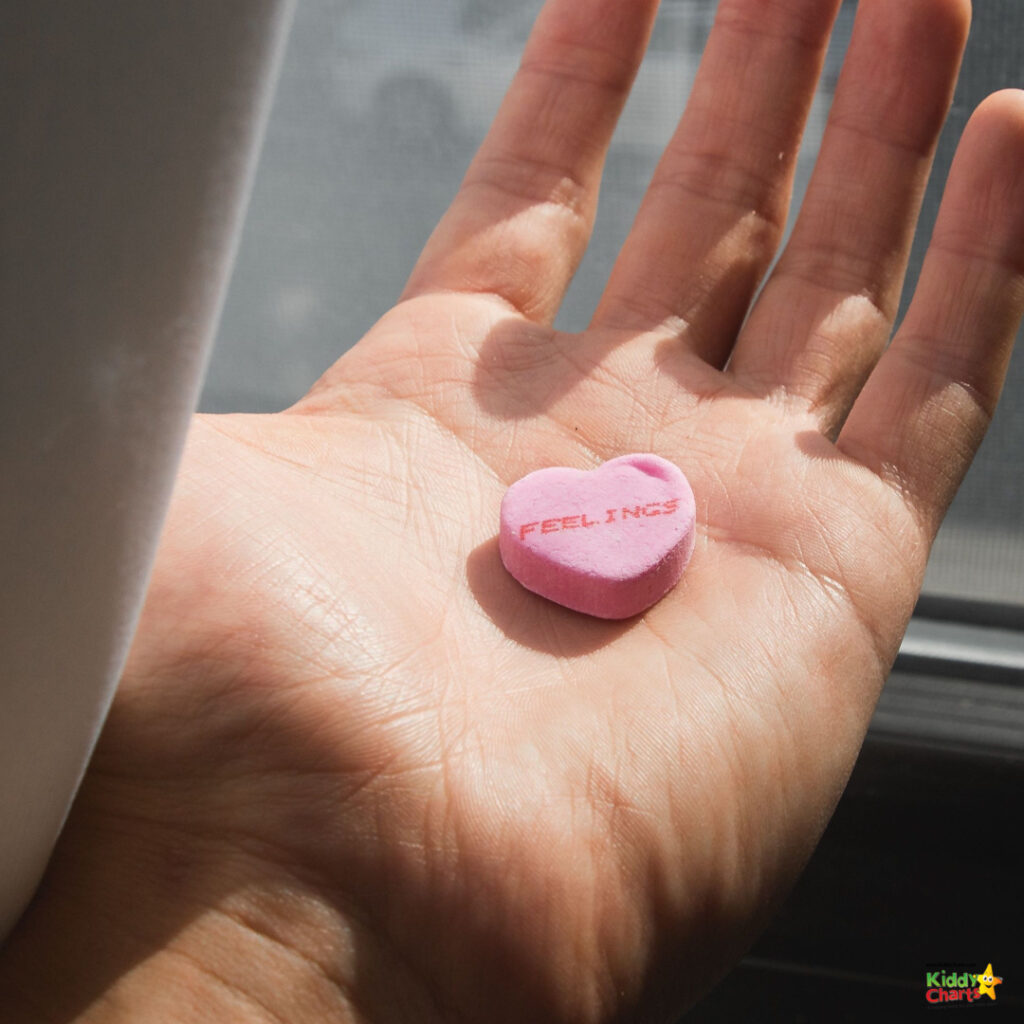 A person's palm is holding a pink heart-shaped candy with the word "FEELINGS" stamped on it, illuminated by natural light near a window.