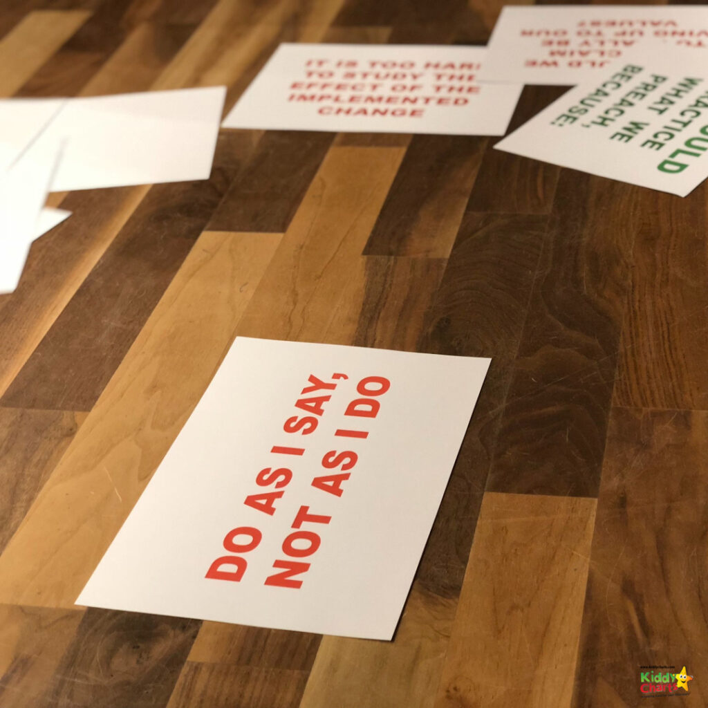 The image shows printed cards with red text scattered on a wooden floor. The visible card reads "DO AS I SAY, NOT AS I DO."