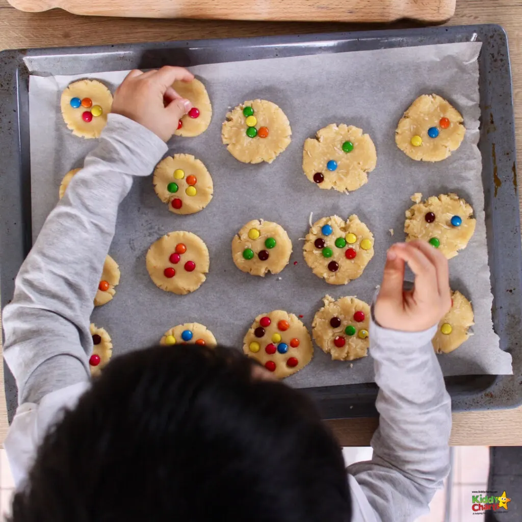 A child is decorating unbaked cookies with colorful candies on a parchment-lined baking sheet, likely preparing them for baking. The setting suggests a home kitchen.