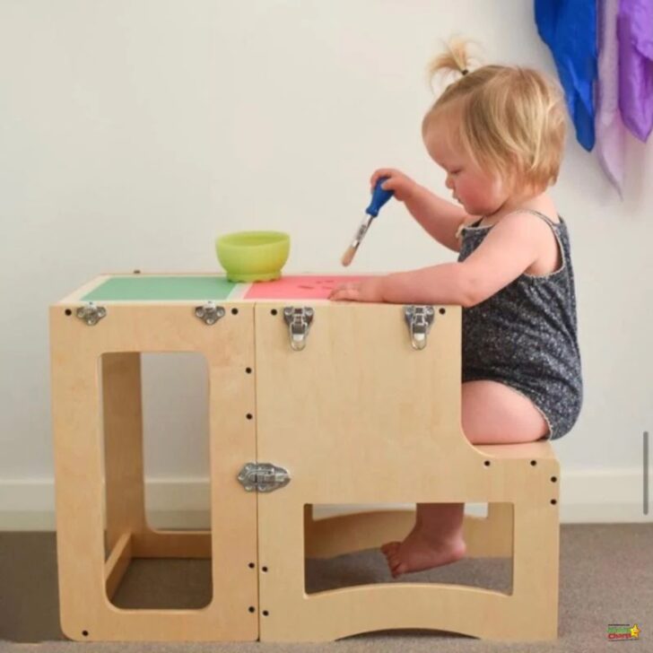 A toddler sits at a wooden multifunctional learning tower, using a brush over a bowl, with papers and focus on a task at hand.