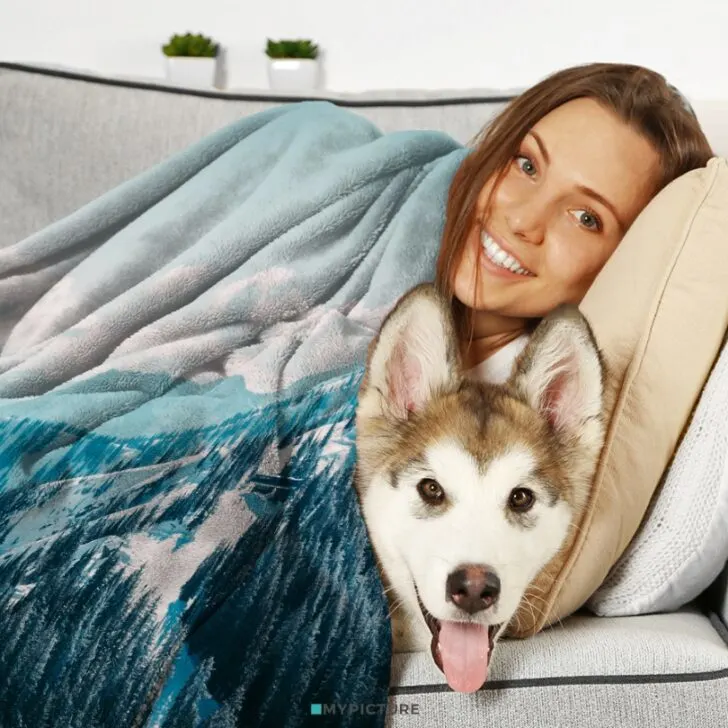 A smiling person and a husky dog snuggled under a blue blanket on a gray couch, with plants in the background. The atmosphere is cozy and happy.