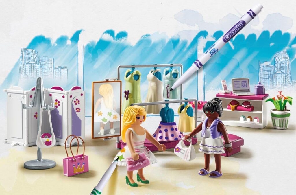 The image shows a colorful playset with two toy figures in a fashion boutique, dresses on racks, accessories, and a Crayola marker emphasizing creativity.