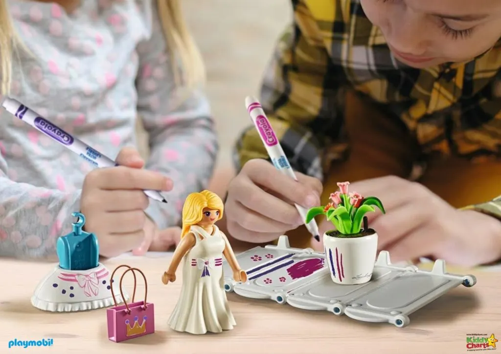 Two children are coloring a Playmobil figure and accessories with markers. A toy flowerpot, handbag, dress, and hangers are visible on the table.
