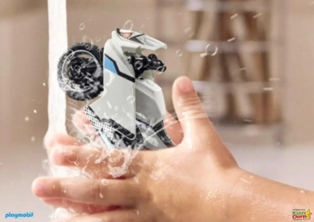 A person's hand is holding a Playmobil toy motorcycle under running water, creating splashes. The image appears playful, with a focus on cleanliness and fun.