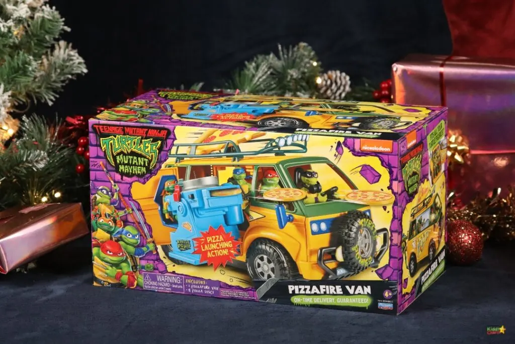 A teenage mutant ninja turtle-themed pizza-launching action vehicle is decorated with a Christmas tree and toy box, and is parked indoors.
