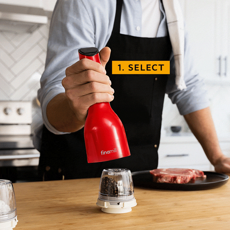 A person in a black apron is holding a red spice grinder above interchangeable pods on a wooden counter, with a steak nearby. The image indicates "1. SELECT".