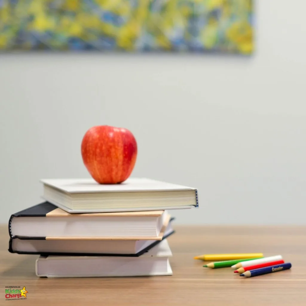 A stack of books on a table with a red apple on top, next to scattered colored pencils, against a blurred background with artwork.