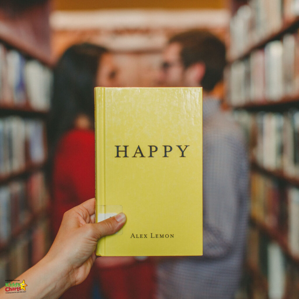 A held book titled "HAPPY" by Alex Lemon is in focus; two blurred people stand in the background within a cozy, bookshelf-lined room.