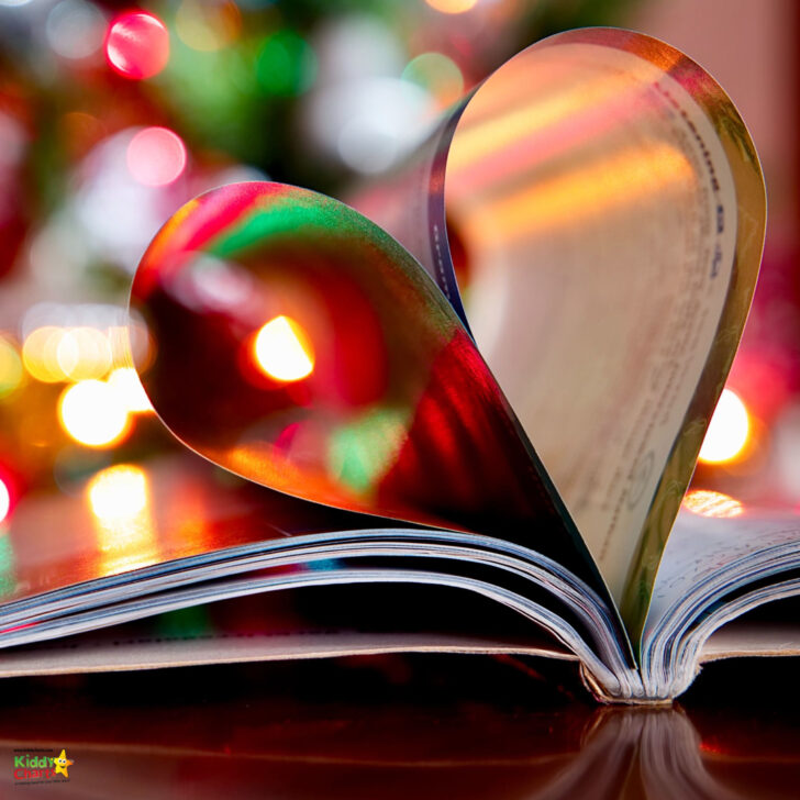 An open book with pages folded into a heart shape sits in the foreground with colorful, defocused lights creating a festive bokeh effect in the background.