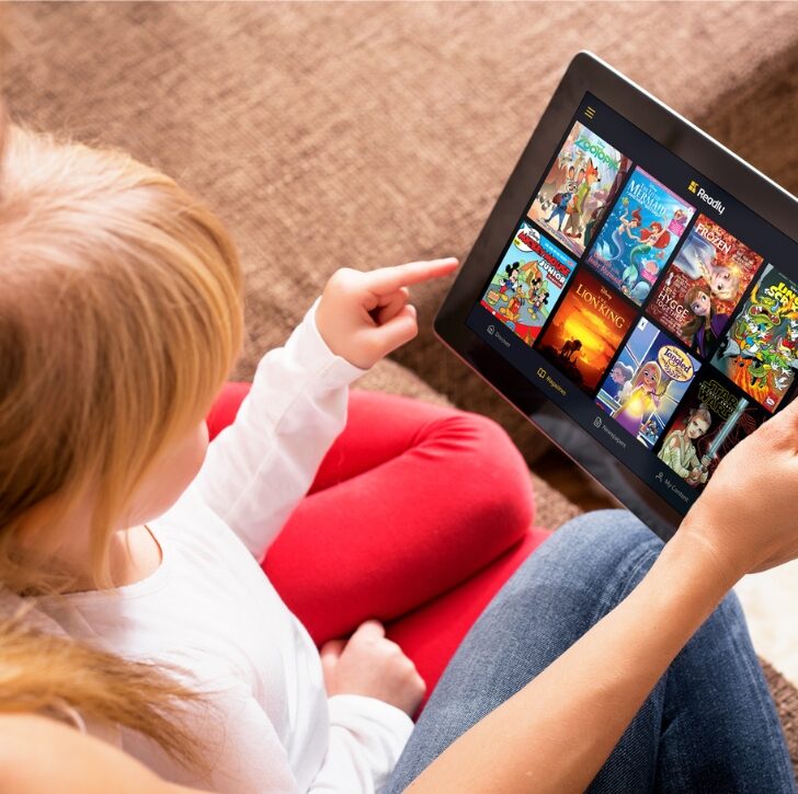A child and a person are looking at a tablet showing a selection of colorful animated movie icons, suggesting they're choosing a film to watch.