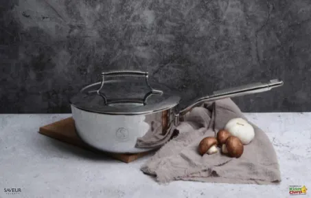 A stainless steel saucepan with a glass lid rests on a wooden board beside mushrooms and a cloth on a textured gray background.