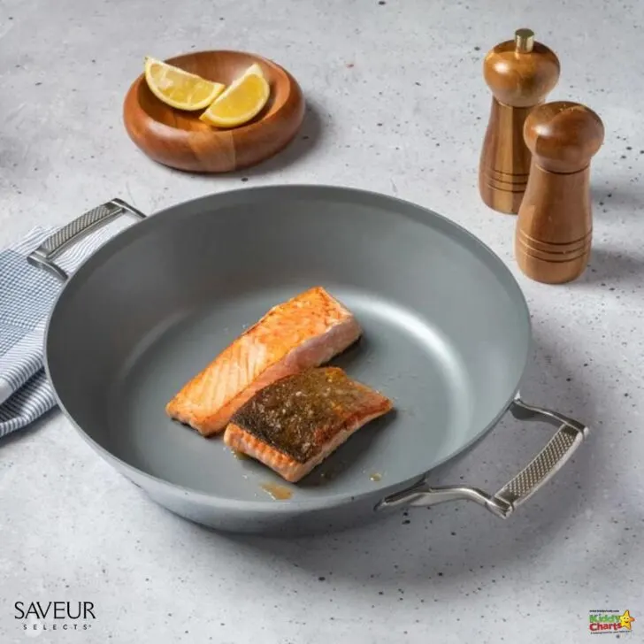 Two cooked salmon fillets rest in a grey non-stick frying pan. Nearby are a salt shaker, pepper grinder, lemon wedges, and a striped napkin.