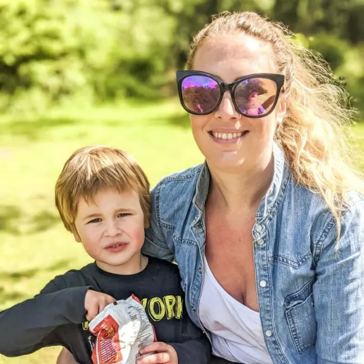 A person with sunglasses and a child are smiling outdoors in a sunny park. Both are casually dressed and seem happy.