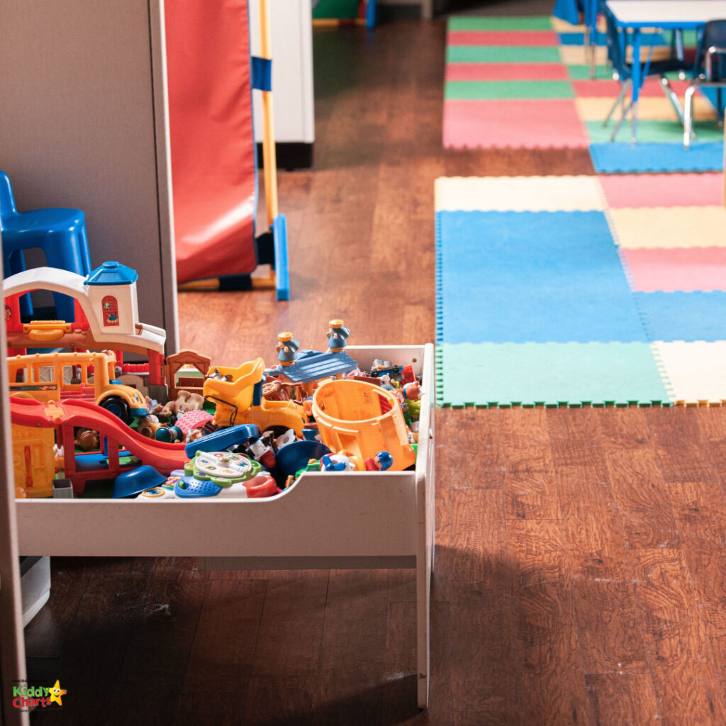 A colorful preschool room with toy-filled shelves, interlocking foam floor mats, child-sized chairs and tables, and educational play areas.