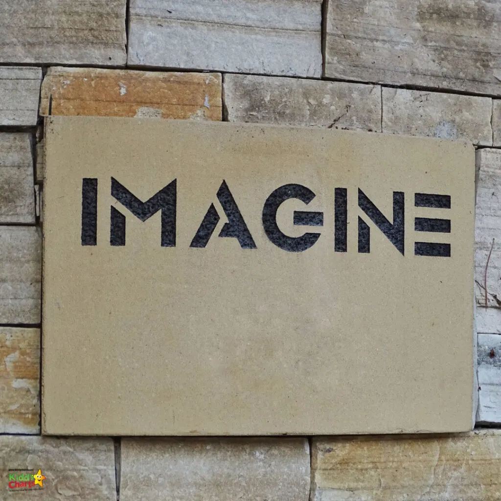 A cardboard sign with the word "IMAGINE" in black letters is mounted on a stone wall. The background shows uneven beige stone bricks.