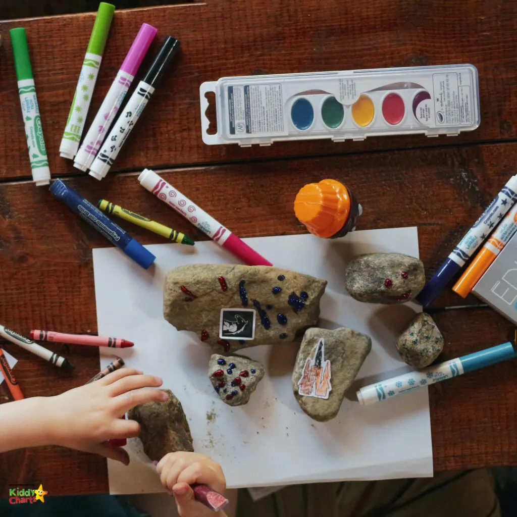 A child's hands are decorating rocks with markers and stickers on a wooden surface, surrounded by colorful art supplies and a watercolor set.
