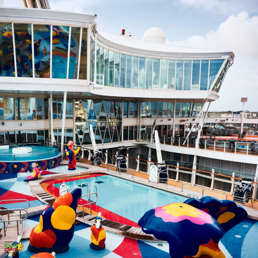The image shows a colorful cruise ship deck with pools, playful sculptures, loungers, and a multi-story glass structure under a blue sky.