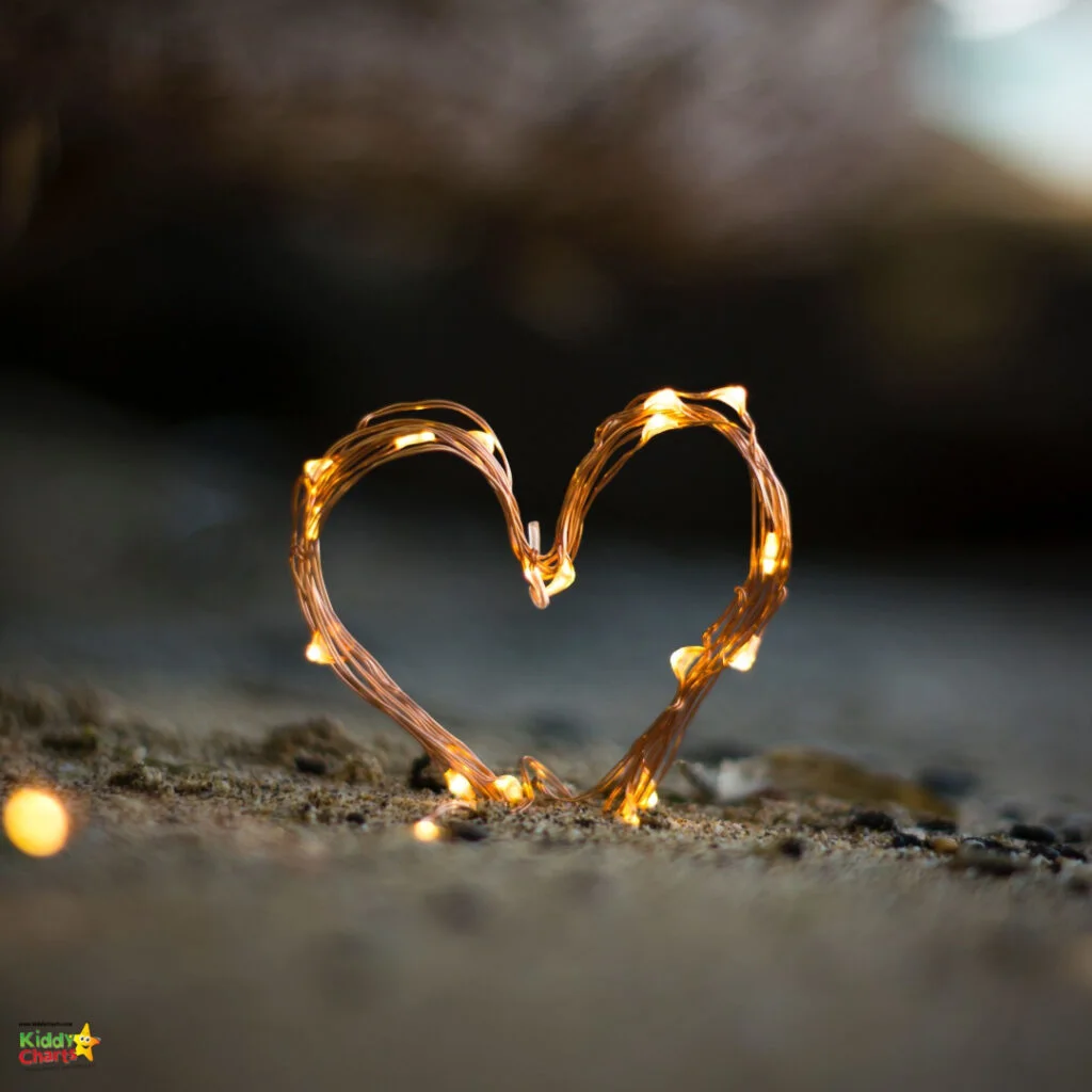 A string of warm, glowing fairy lights twisted into a heart shape on a sandy surface with a soft-focus background.