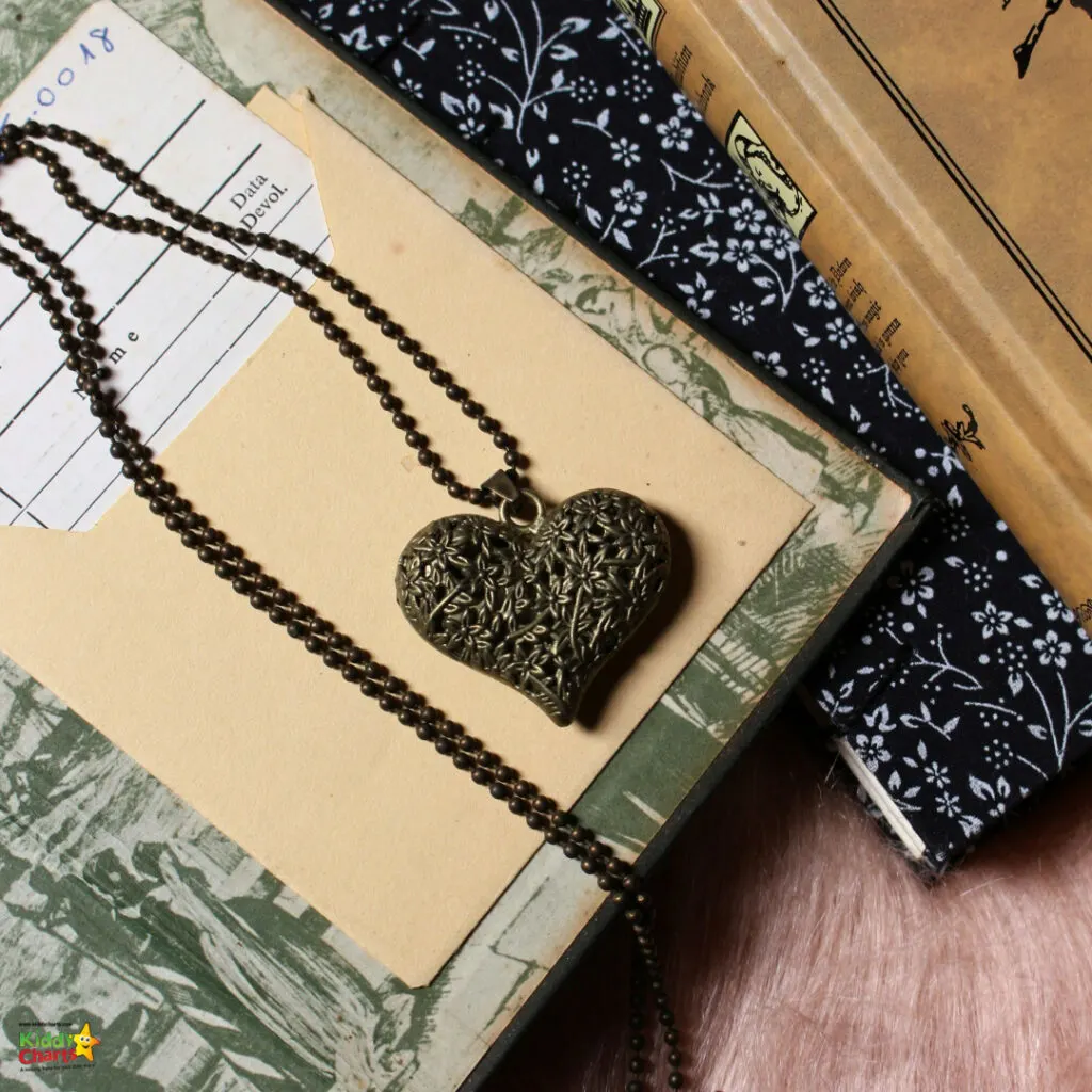 This image features a heart-shaped pendant with intricate patterns on a beaded chain, laid across a manila folder and vintage books with floral fabric.