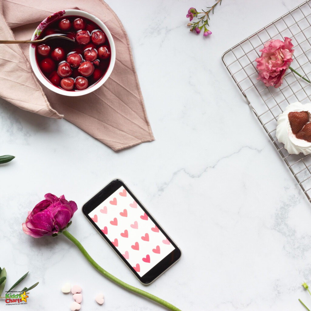 A flat lay composition with cherries in a bowl, a smartphone with a heart-themed wallpaper, fresh flowers, a cake, and a cooling rack on a marble surface.