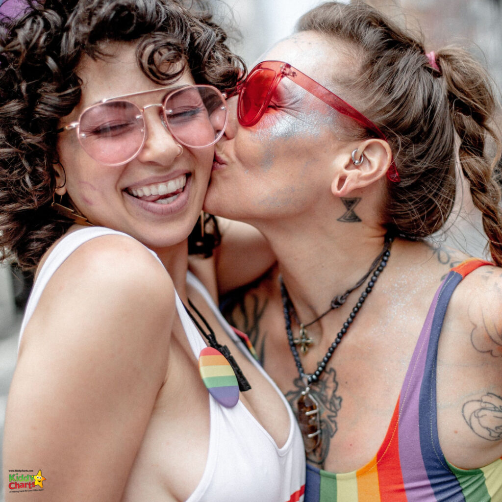 Two joyful people embrace and share a kiss on the cheek. They wear colorful attire, sunglasses, and have tattoos, exuding a sense of celebration and happiness.